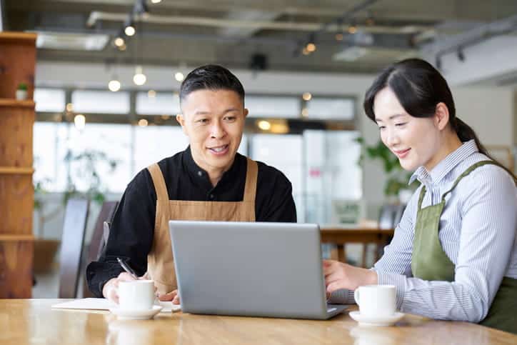 Two restaurant owners sit at a table and discuss their restaurant marketing strategies while looking at a laptop.